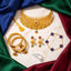 A styled flat-lay of traditional Indian gold jewelry on a white marble slab surrounded by draped blue, green, and burgundy satin. The jewelry set includes a gold choker necklace with a tassel, a stack of gold bangles with green and red enamel details, a pair of gold earrings with floral tops, a gold bracelet with blue enamel clover motifs, and a silver ring with a clear gemstone.
