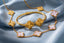 Gold jewelry set featuring a bracelet and necklace with clover-shaped links, some inlaid with pale pink shell, displayed on draped light blue fabric.