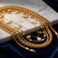 A layered set of gold-tone necklaces, including a chunky curb chain, a braided chain, delicate chain links, and several floral charms, some accented with small white pearls, displayed on a white marble slab over a dark blue velvet background.