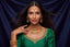 A woman with long dark hair models an ornate gold jewelry set, including a necklace, earrings, ring, and bangles, paired with a green embroidered traditional top against a dark blue satin background.