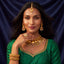 A woman with long dark hair models an ornate gold jewelry set, including a necklace, earrings, ring, and bangles, paired with a green embroidered traditional top against a dark blue satin background.