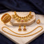 Gold-toned traditional jewelry set arranged on a white marble slab with dark blue fabric in the background. The set includes a necklace, matching earrings, a bracelet, and a ring, decorated with gold, white pearls, and pink stone accents.